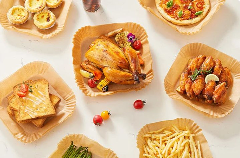Vista cenital de varios platos de comida preparados en la freidora de aire (pollo asado, alitas, pizza, tostadas, patatas fritas) servidos en moldes de papel de horno desechables sobre una mesa blanca